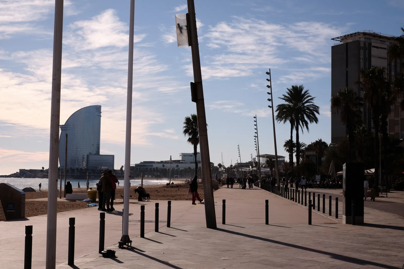 Barcelona waterfront promenade at golden hour, with the W Hotel sail-shaped silhouette in the background, palm trees lining the boardwalk, and pedestrians walking along the shore.