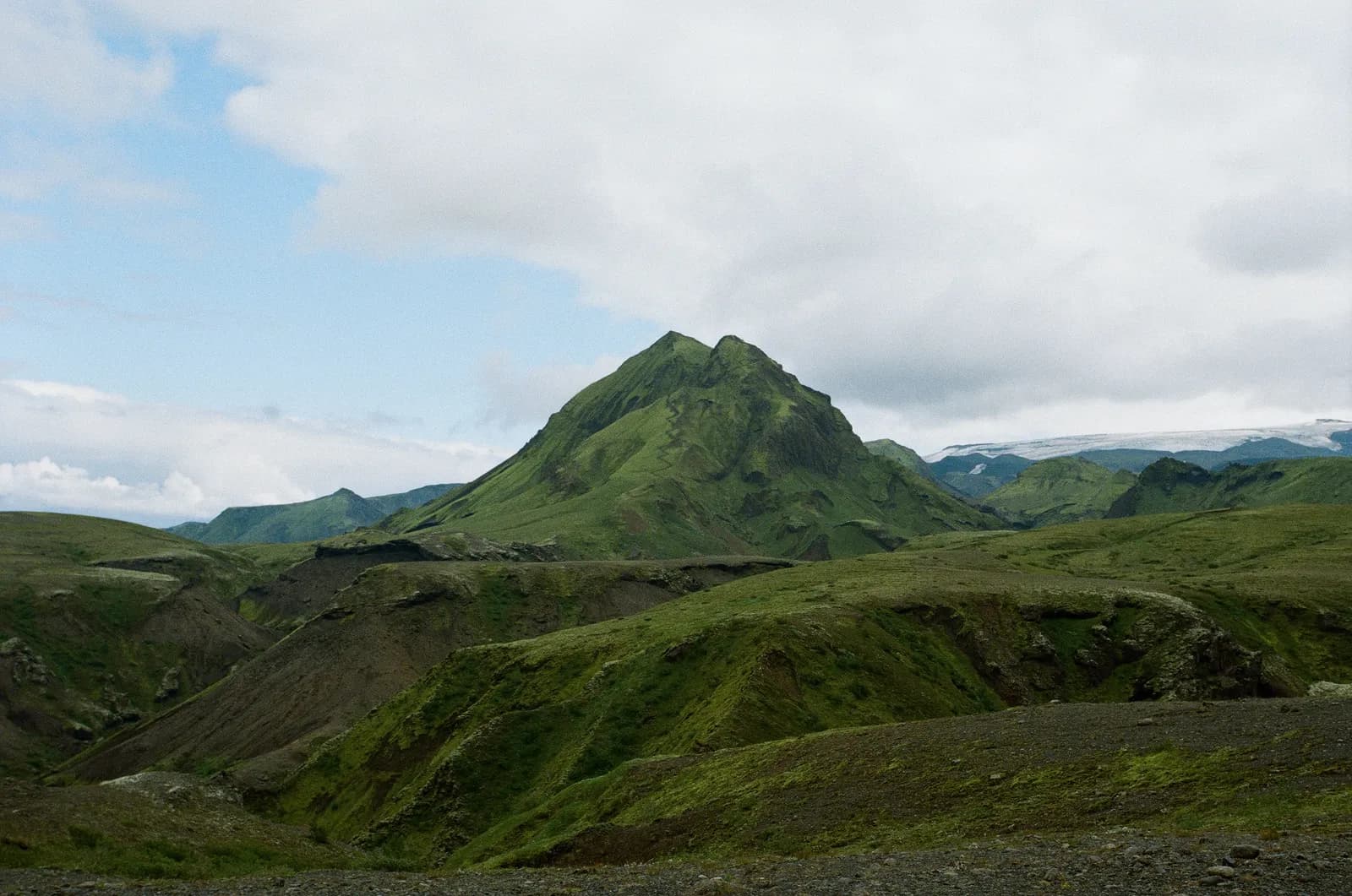 A single conical green mountain rising from a sea of lower ridges and dark gravel paths in Iceland. The peak holds steady at the centre of the frame while the layered terrain around it shifts away. The visual analogue to a brand anchored at the top of an AI response while the supporting cast of cited URLs churns underneath.