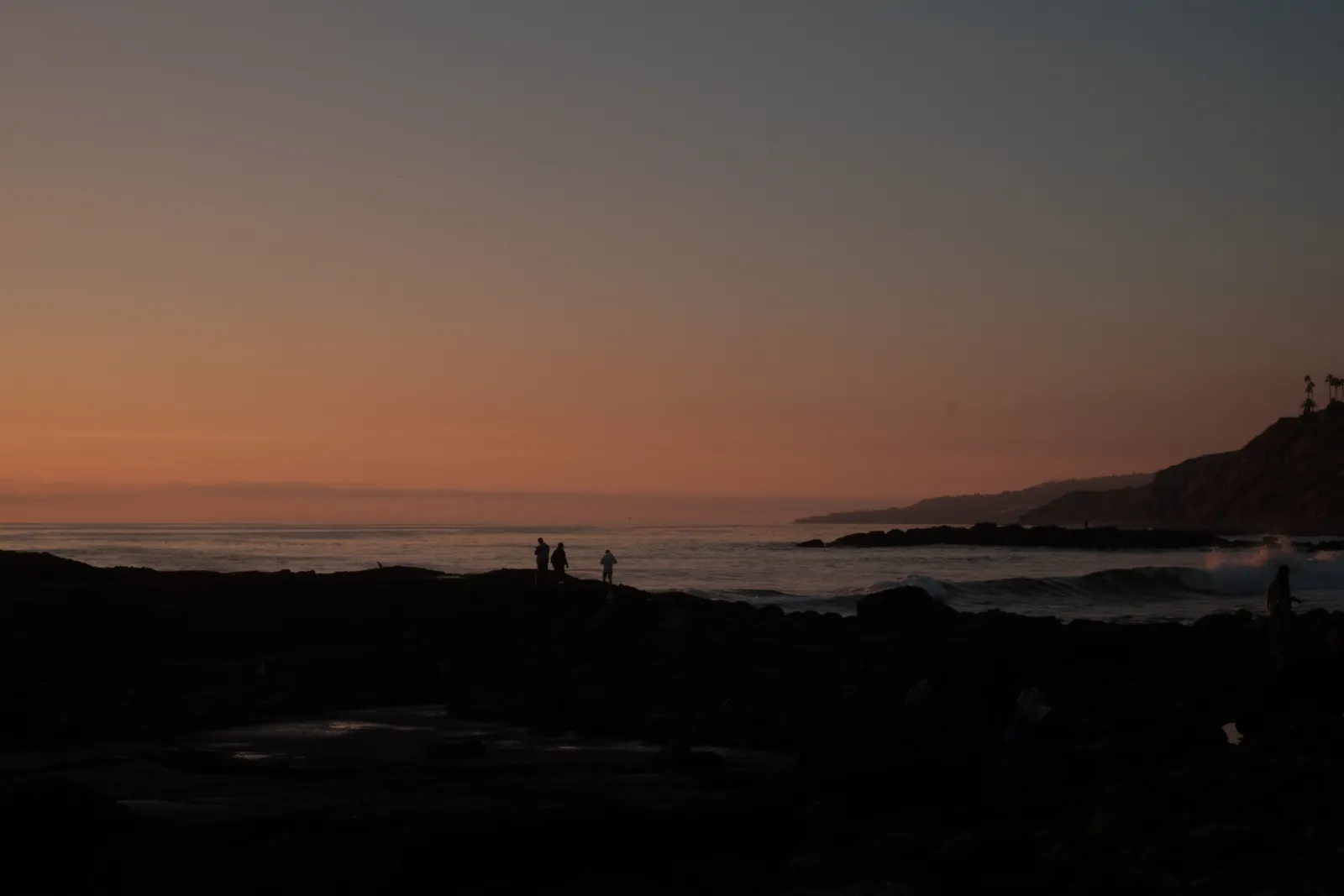 Silhouettes of people standing on a rocky coastline at sunset, looking out at the ocean. The warm orange horizon fades into darkness below, a visual metaphor for content creators watching their work recede into the background of AI answers while competitors stand in the spotlight.
